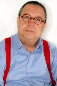 A headshot of Eduardo Aguado López, a man wearing red circular glasses, a blue button up shirt, and red suspenders.