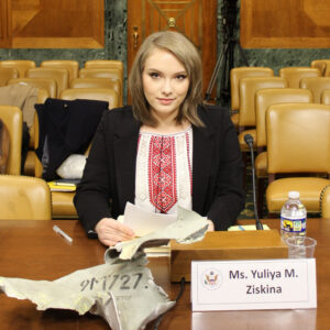 Woman sitting at table in U.S. Helsinki Commission hearing, showcasing shrapnel from Russian missile strike in Ukraine.